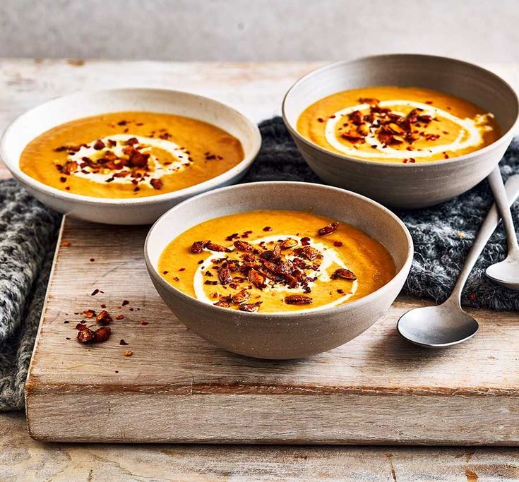 three bowls of soup on a cutting board with silver spoons and utensils