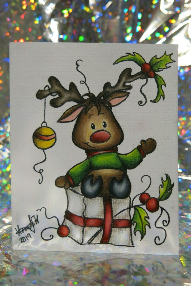 a christmas card with a reindeer sitting on top of a present box and holding an ornament