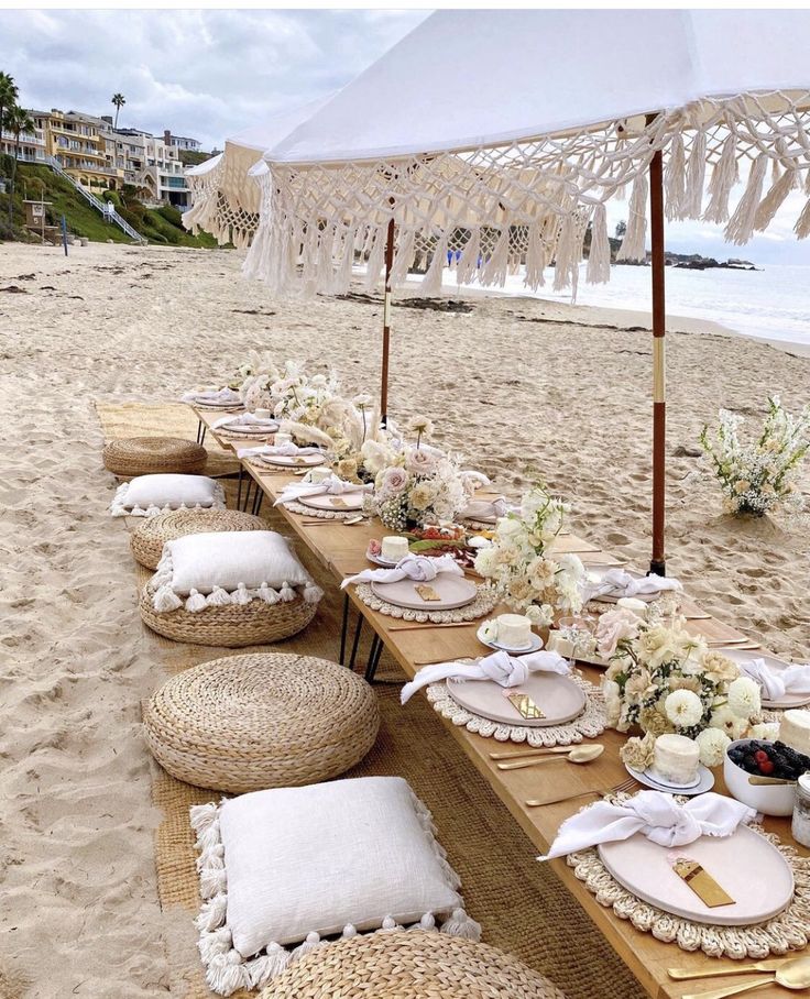 a long table set up on the beach with white flowers and place settings under an umbrella