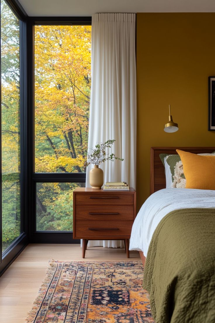 a bedroom with a bed, dresser and large window overlooking trees in the fall season