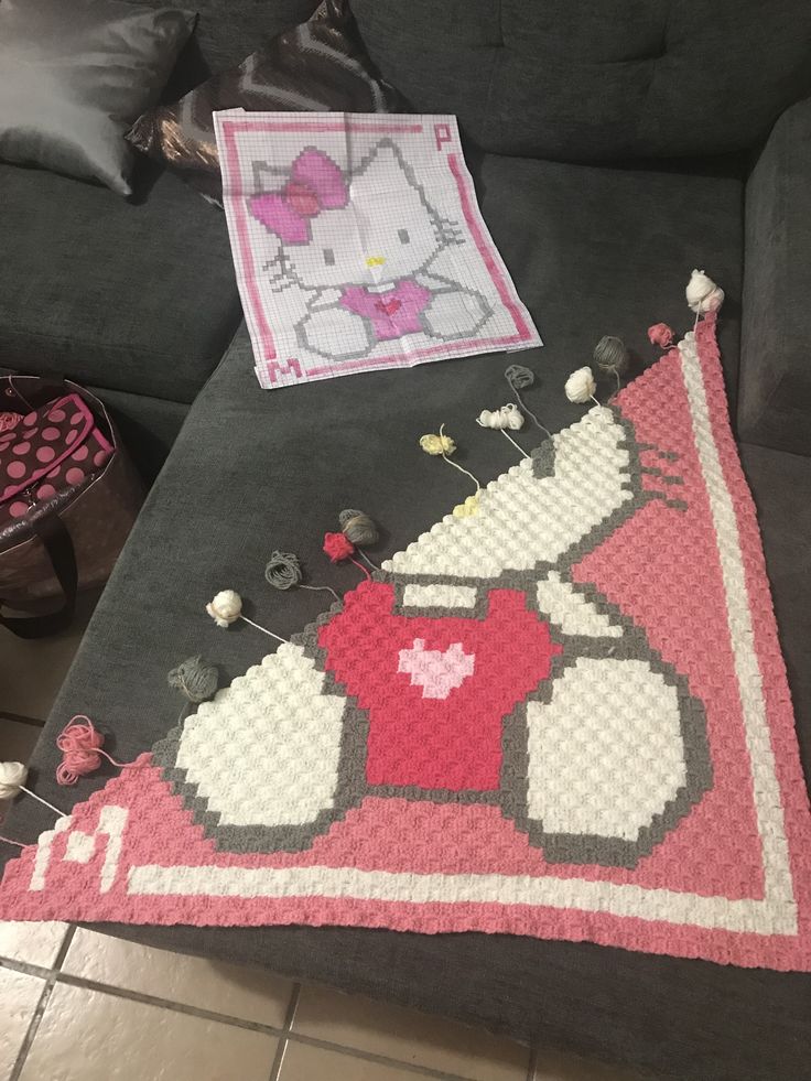 a hello kitty rug sitting on top of a gray couch next to a pink and white pillow