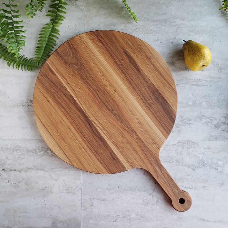 a cutting board with a wooden handle next to a green plant and an apple on the table