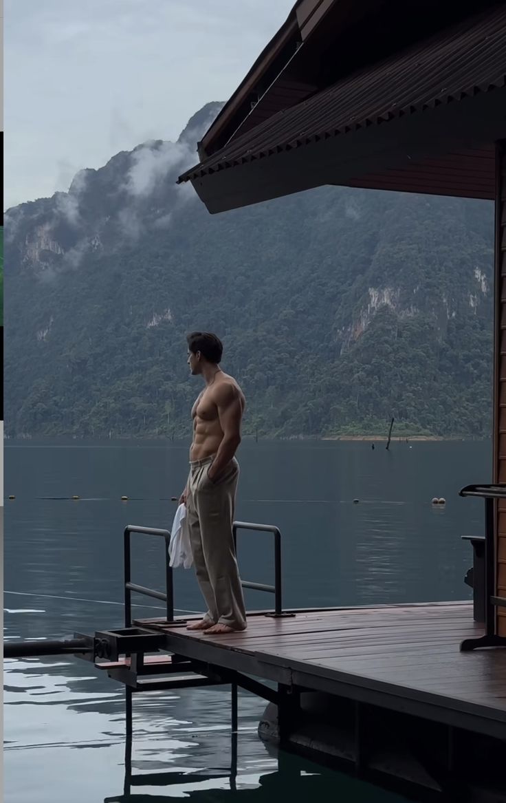 a shirtless man standing on a dock next to a body of water with mountains in the background