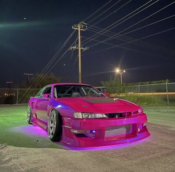 a pink car parked in the middle of a parking lot at night with its lights on