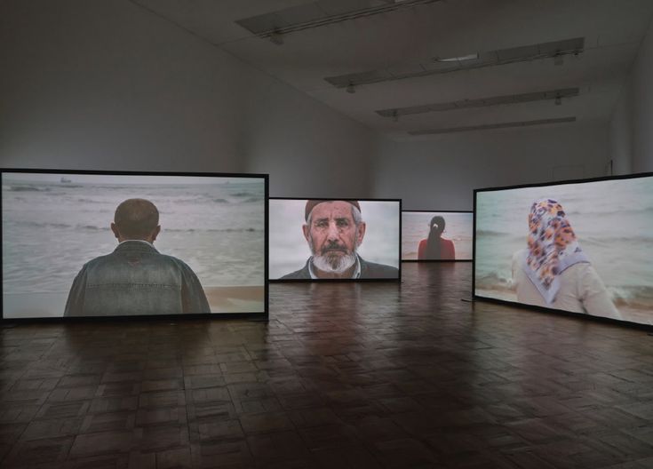 an art gallery with four large screens displaying people's faces and images on them