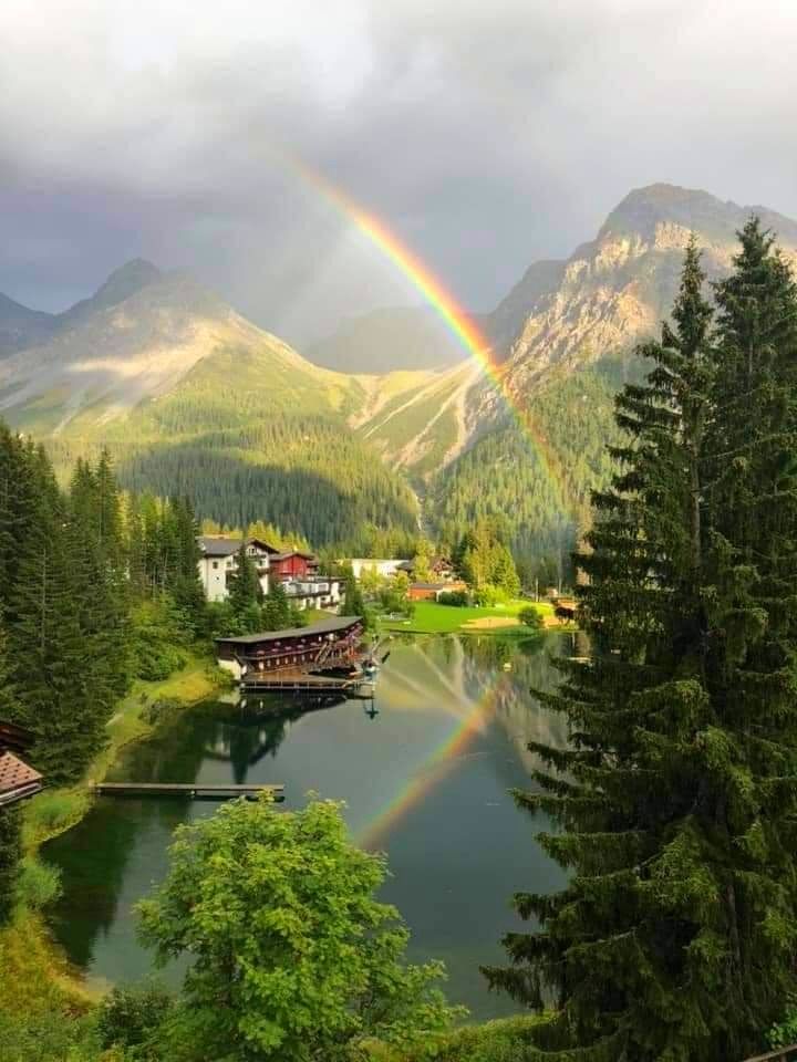 a rainbow in the sky over a lake with houses on it and mountains in the background