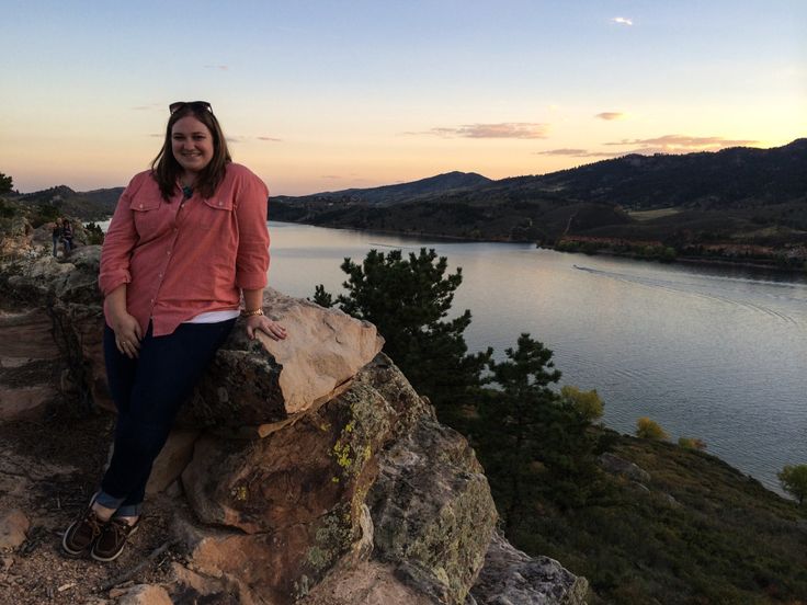 a woman sitting on top of a rock next to a body of water at sunset