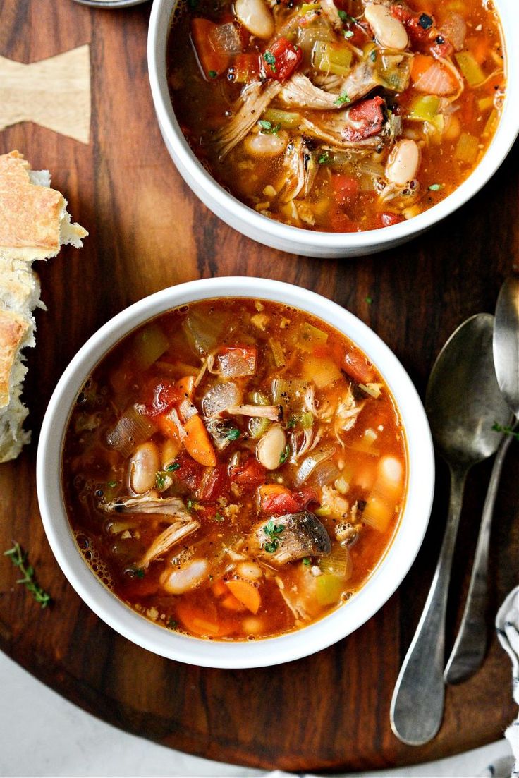 two bowls of vegetable soup on a wooden table with spoons and bread in the background