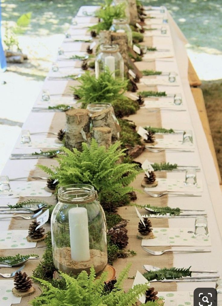 a long table is set with pine cones, ferns and candles for an outdoor dinner