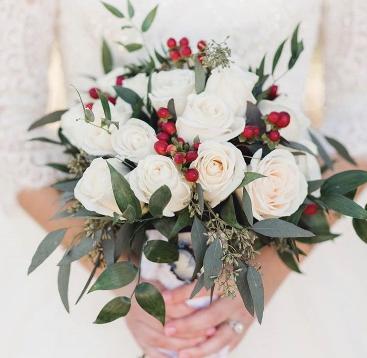 a bride holding a bouquet of white roses and greenery in her hands with red berries on the stems