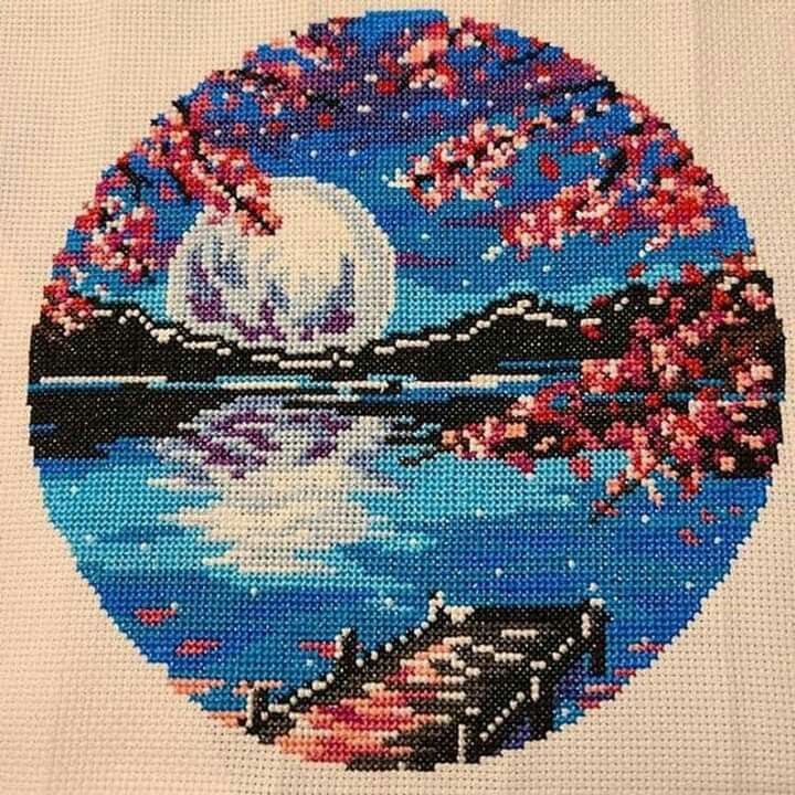 a cross - stitch picture of a lake with cherry blossoms in the foreground and a full moon in the background