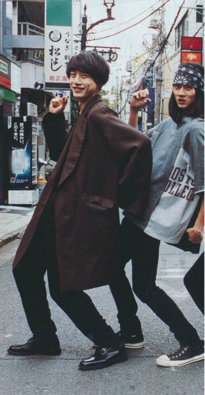 two young men walking down the street with one pointing at something in his hand and another holding an umbrella