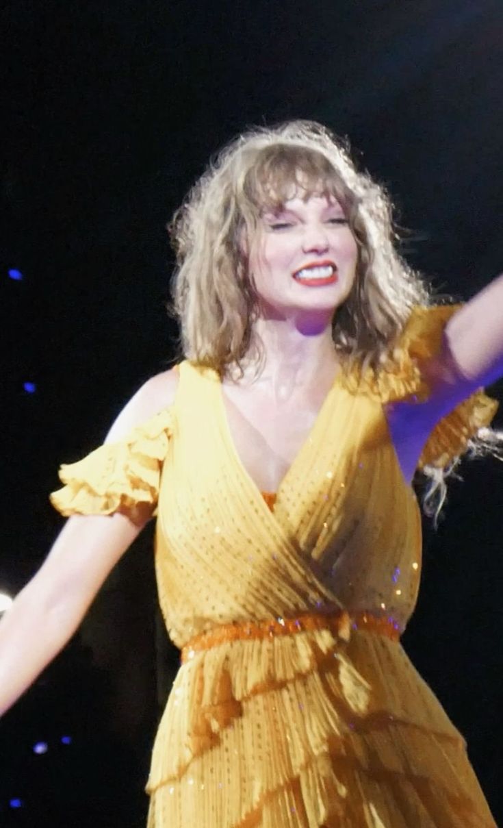 a woman in a yellow dress on stage with her arms out and one hand raised