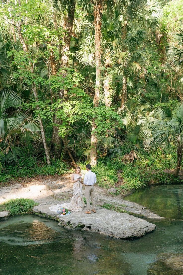 a man and woman standing on a rock in the middle of a river surrounded by trees