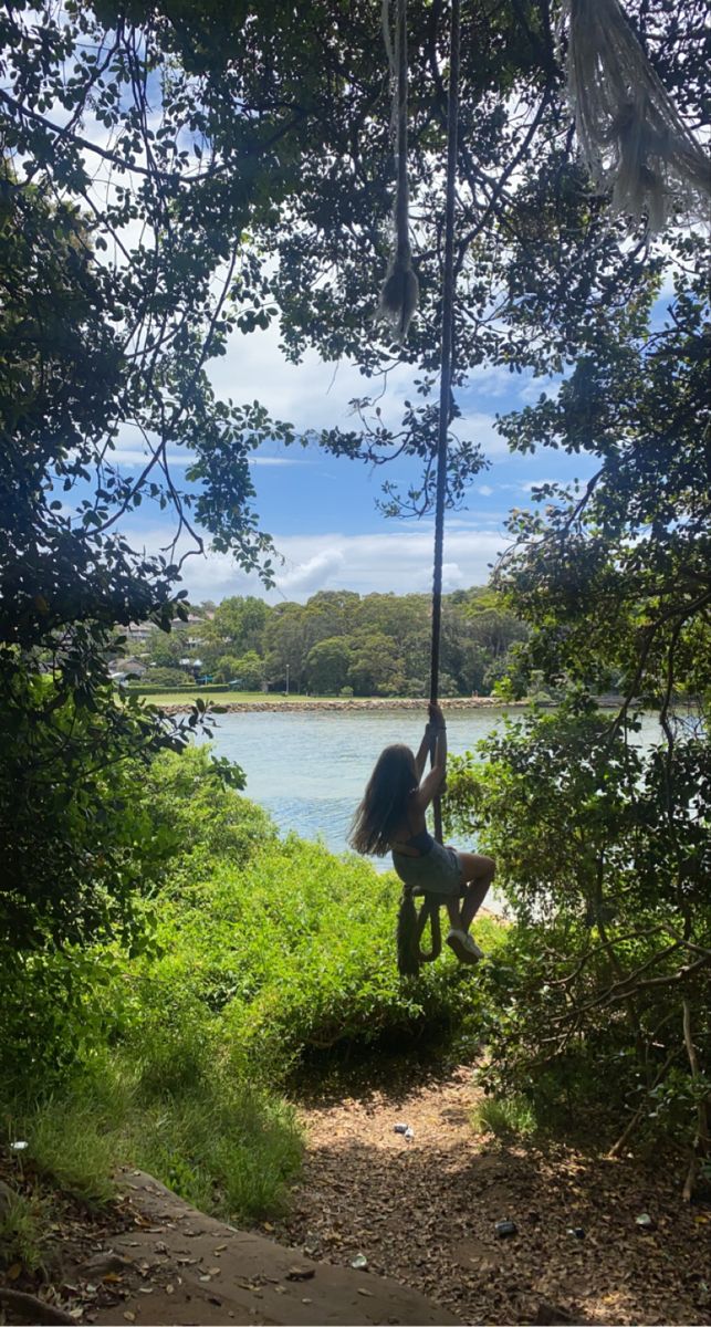 a girl is swinging on a rope in the woods next to a body of water