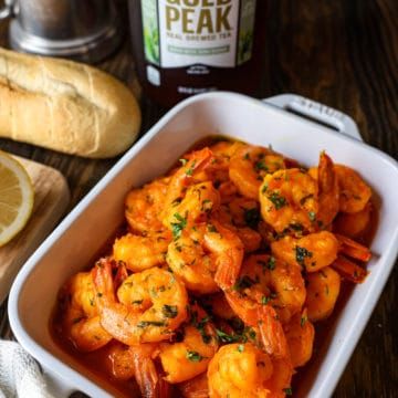 a white dish filled with cooked shrimp next to bread and lemon wedges on a wooden table