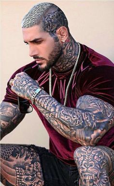 a man with tattoos on his arms and chest sitting down in front of a white wall