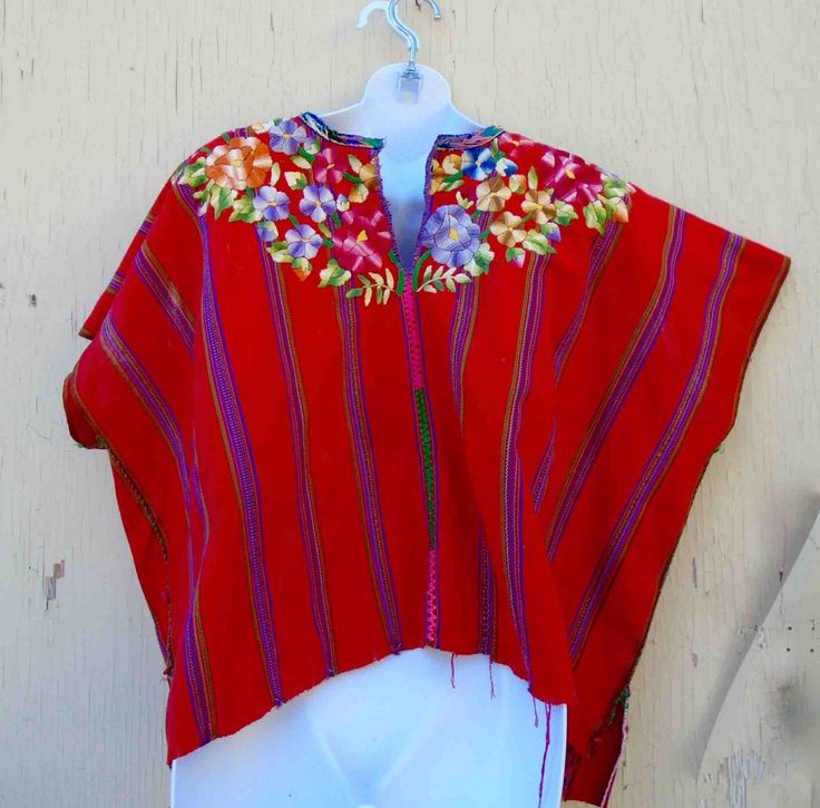 "A wide all cotton hand made vintage blouse or huipil that is brilliant bright red all over, with a border of hand-embroidered flowers and leaves at neck area of blouse, both front and back. Sleeves are short, wide at shoulders, has ten vertical embroidered rainbow ( multicolored) travel down top, each speed 2\" apart. Center line has a band of blue, pink, and green embroidery running down to bottom. Top is hand-sewn up each side with bright green thread, and bottom hem is a rough extra panel as Bohemian Red Blouse With Traditional Patterns, Red Folk Style Tops For Festive Occasion, Traditional Red Blouse With Traditional Patterns, Traditional Red Tops For Festival, Red Blouse With Traditional Patterns For Festivals, Red Folk Style Tops For Festivals, Traditional Red Blouse For Summer, Traditional Red Blouse With Embroidered Border, Red Bohemian Blouse With Traditional Patterns