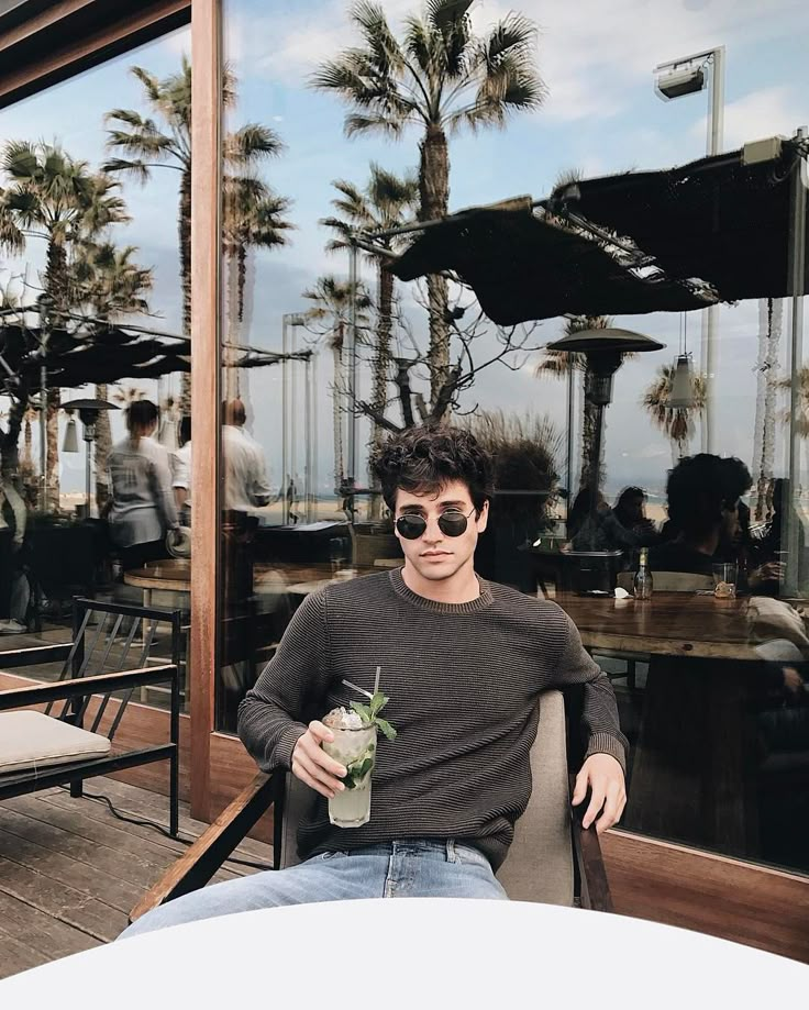 a man sitting at a table with a drink in his hand and palm trees behind him