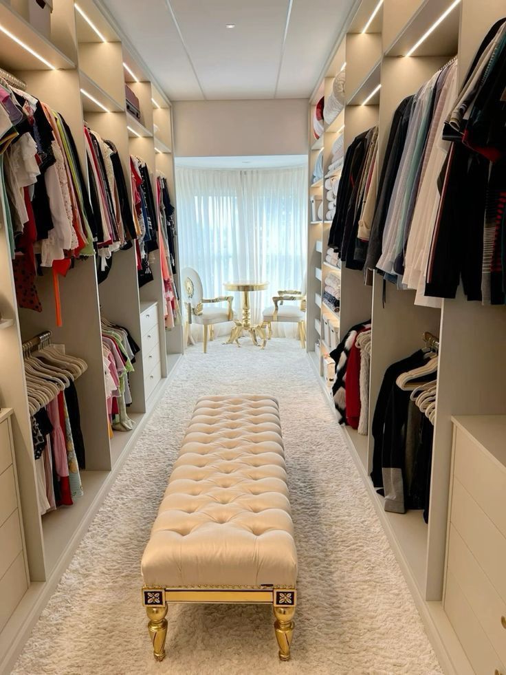 a walk in closet with a bench and clothes on shelves next to the door that is open