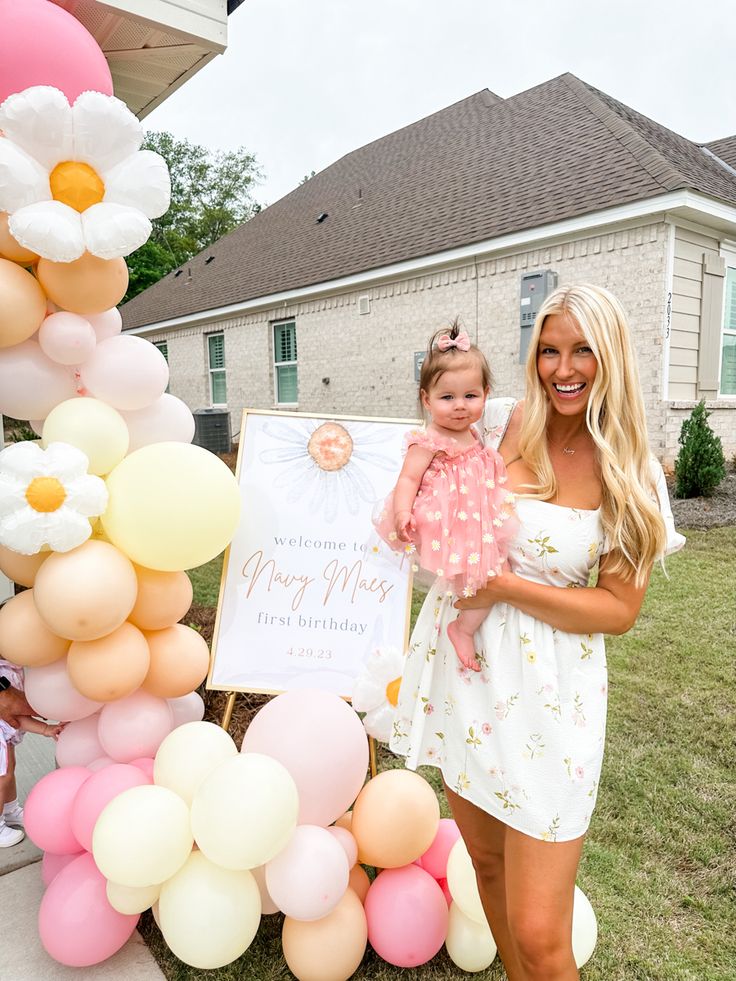 a woman holding a baby standing next to a sign with balloons and flowers on it