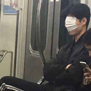 two people sitting on a train with masks covering their faces while looking at their cell phones