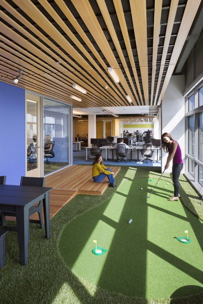 two people playing miniature golf in an office setting with large windows and grass on the floor