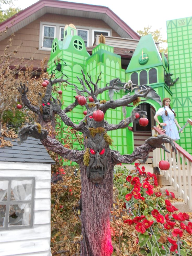 a fake tree with apples on it in front of a green house and red flowers