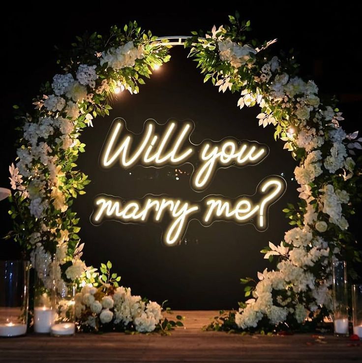 a lighted sign that says will you marry me? surrounded by white flowers and greenery