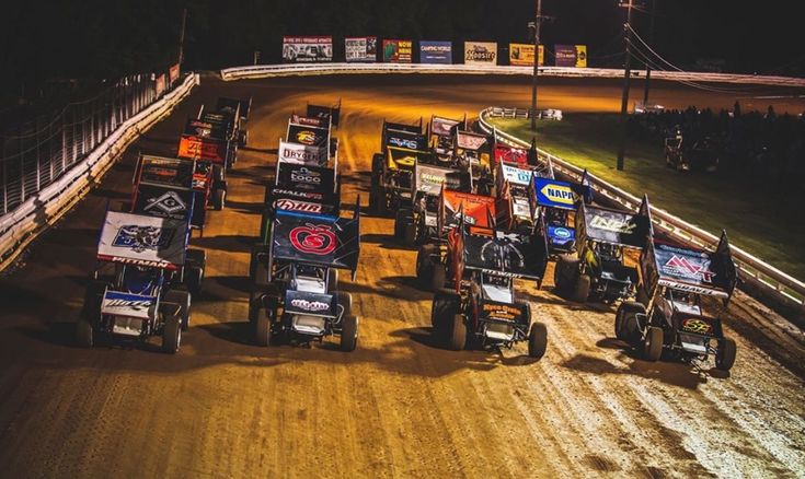 a group of dirt racing cars driving down a race track at night with the lights on