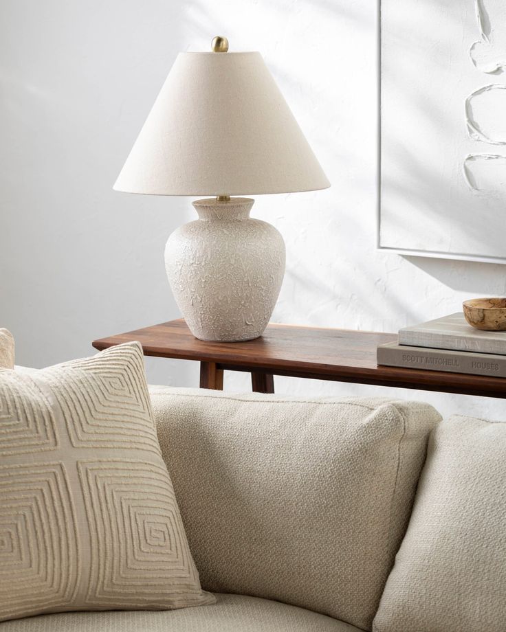 a white lamp sitting on top of a wooden table next to a couch with pillows