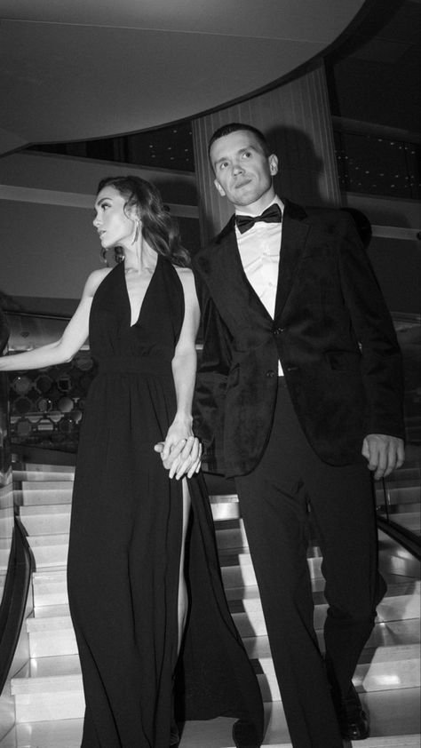 black and white photo of two people in formal wear walking down stairs together, one holding the hand of the other's hand