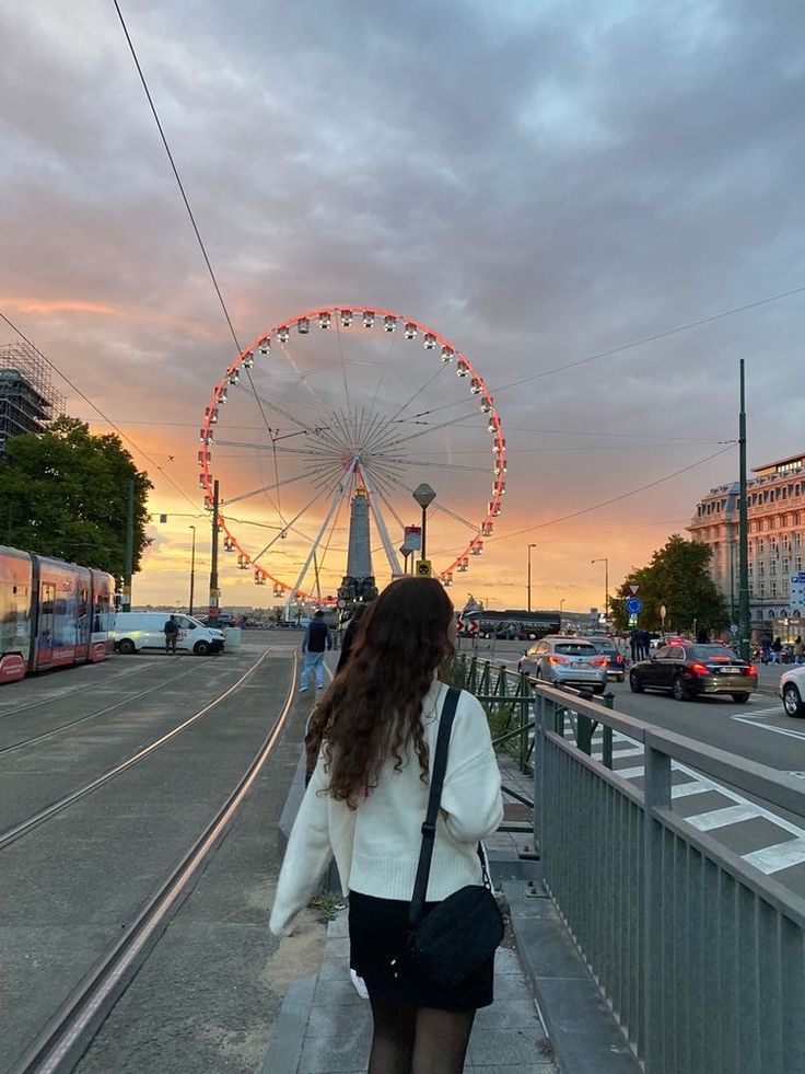 a woman is walking down the sidewalk near a ferris wheel at sunset with cars and buses in the background