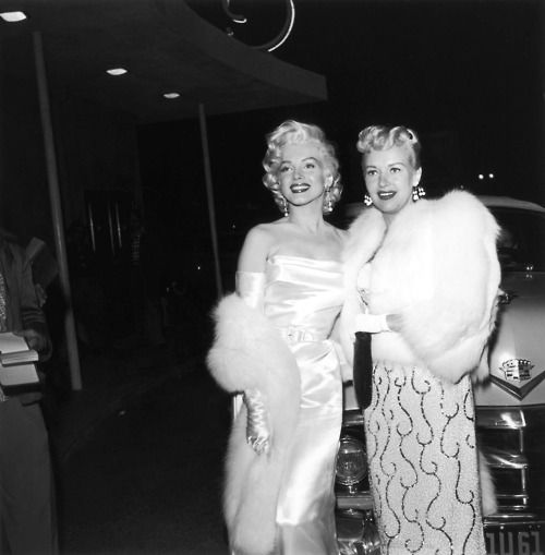 two women standing next to each other in front of a car wearing fur stoles