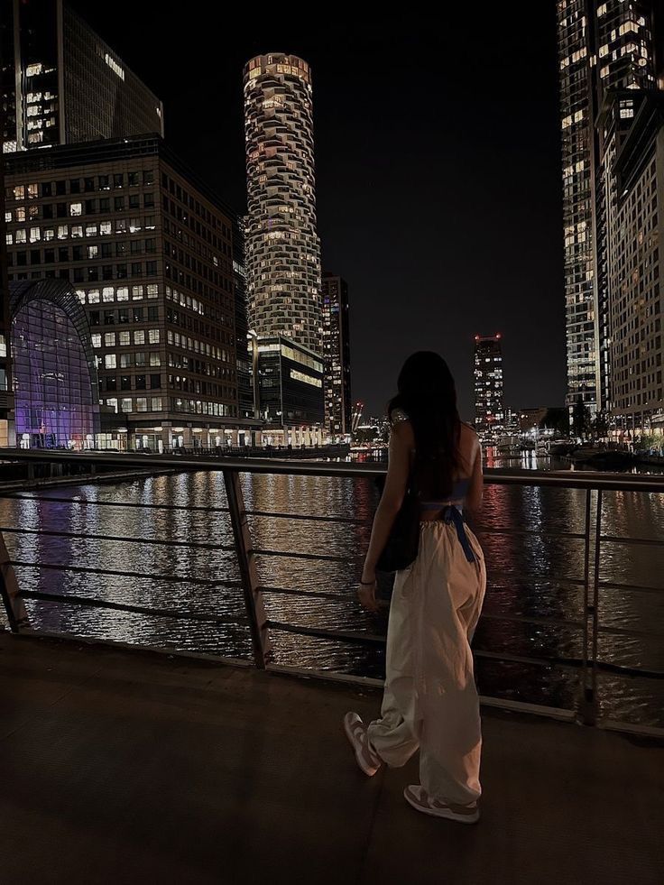 a woman standing on the edge of a bridge looking out over water at night time