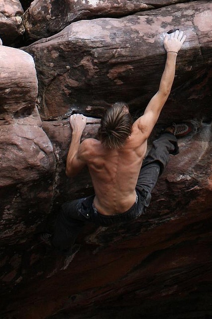 a shirtless man climbing up the side of a rock face with his hands in the air