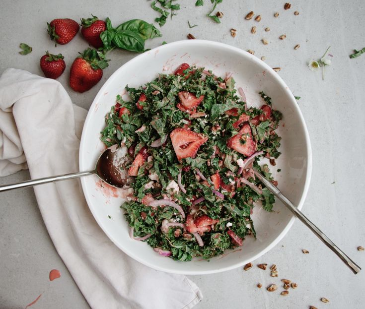 a white bowl filled with salad and strawberries on top of a table next to two spoons