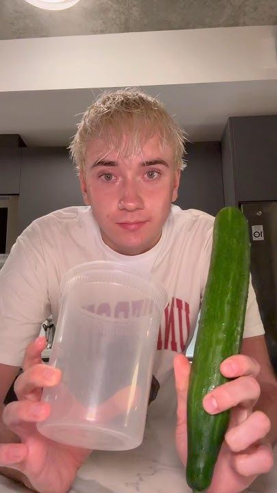 a young man holding a cucumber in a plastic container while sitting at a kitchen counter