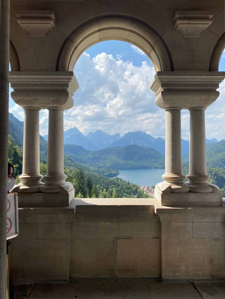 Tan arch with two small columns on each side of it columns are on a base that goes connects to both columns. View is blue sky lots of pretty white clouds, mountains, small view of a lake, and lots of green trees. Greek Castle, Water Wall Fountain, Fake Window, Neuschwanstein Castle, Fantasy Castle, Through The Window, Beautiful Places To Visit, Fantasy Landscape, Amazing Nature