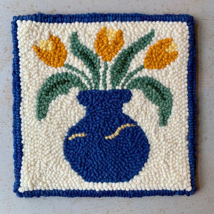 a blue vase with yellow flowers in it on a white and blue square rug that has crocheted border