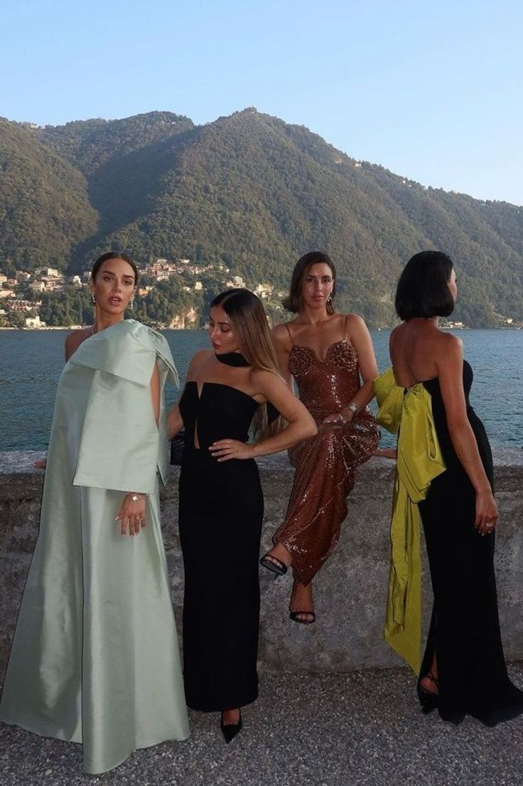 four women standing next to each other in front of the water and mountains with hills behind them