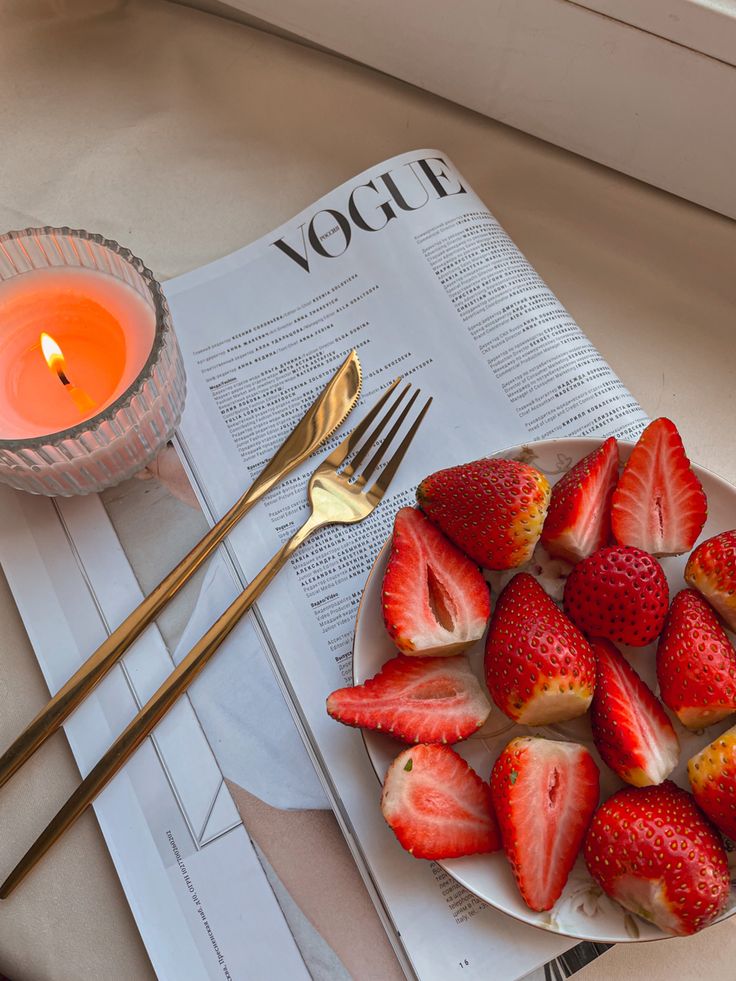 strawberries are on a plate next to a candle and newspaper with utensils