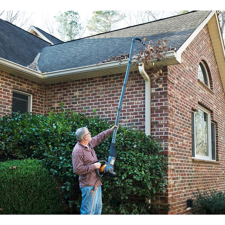 a man using a power washer to clean the roof of a brick house with shrubbery behind him