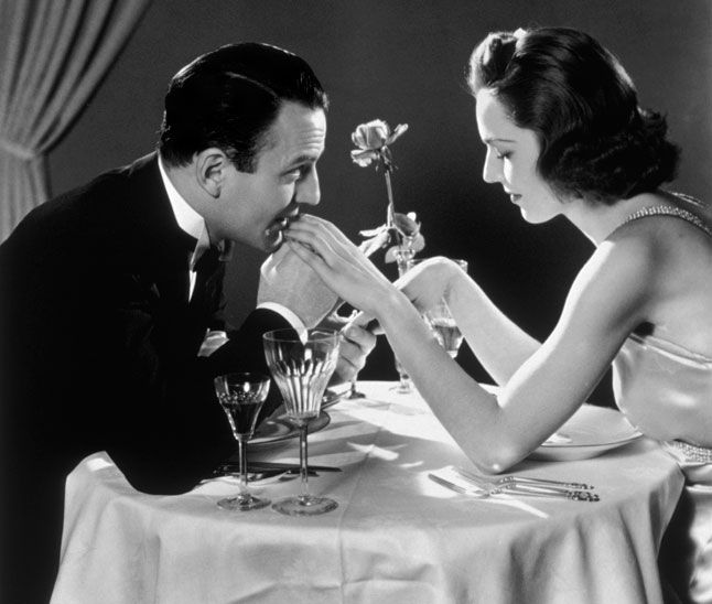 a man and woman sitting at a table with flowers in their hands, one holding the other's hand