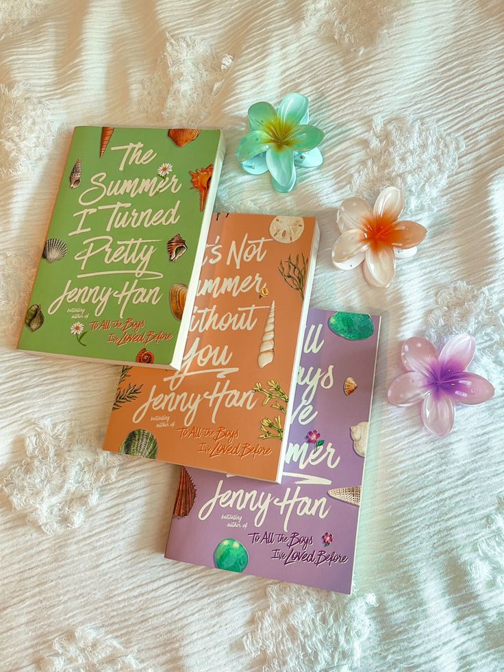 three books laying on top of a white bed next to flowers and an orange flower