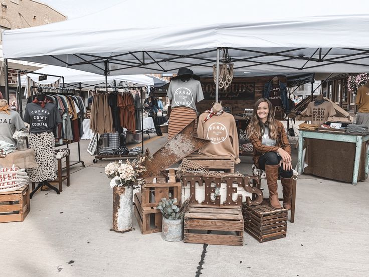 a woman sitting in front of a display of clothes and other items at an outdoor market