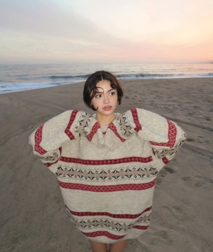 a woman standing on top of a sandy beach next to the ocean wearing a sweater