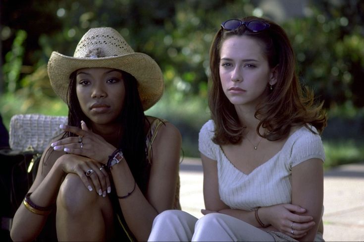 two women sitting next to each other wearing hats and bracelets on the ground in front of trees