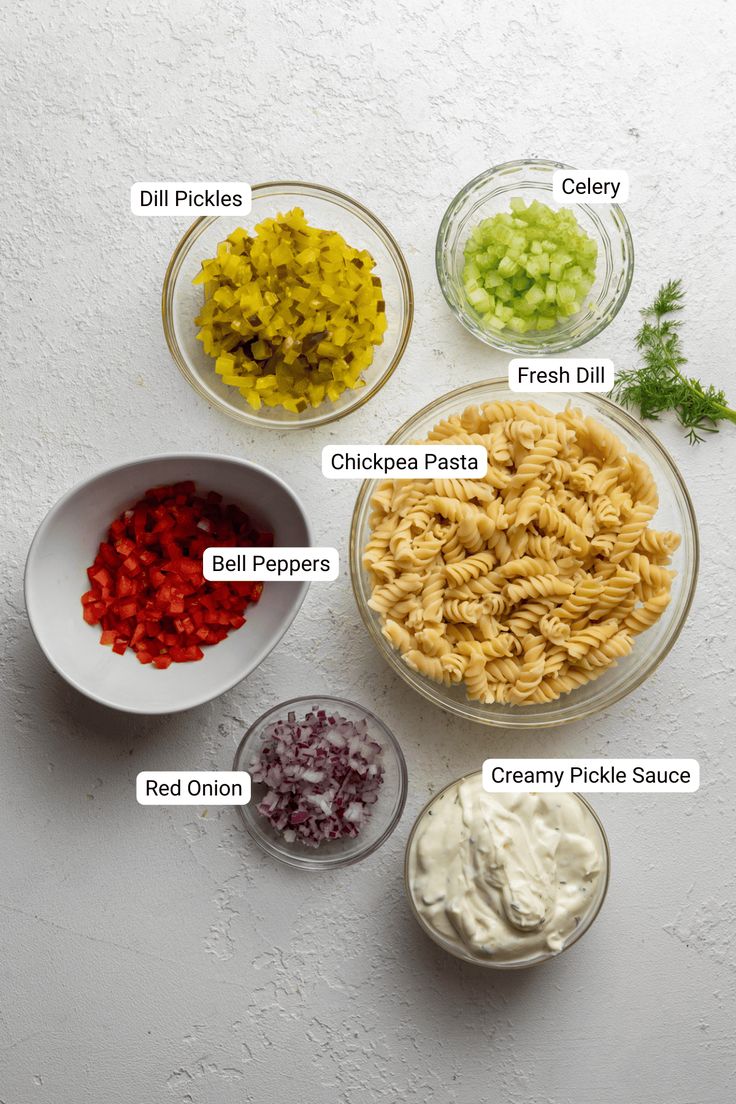 the ingredients for this pasta dish are shown in bowls on a white surface, including onions, celery, red onion, and green pickles