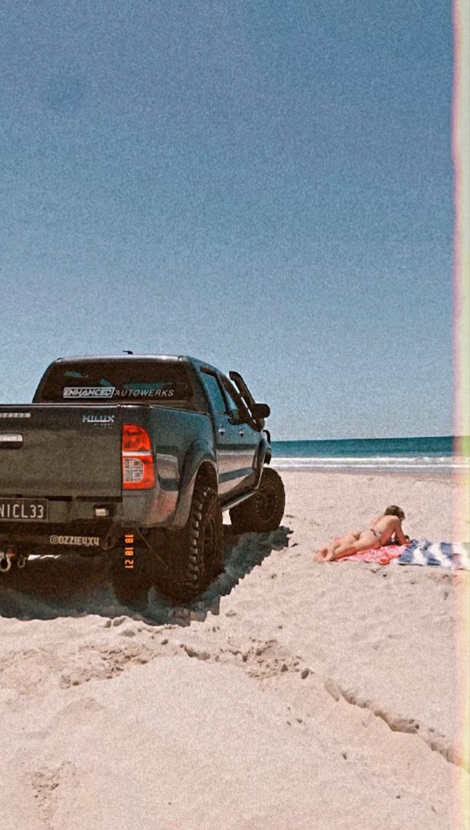 a pickup truck parked on the beach next to a person laying in the sand